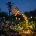 Fairy Watering Can Light glowing at dusk in a garden flower bed, warm light pouring from the spout